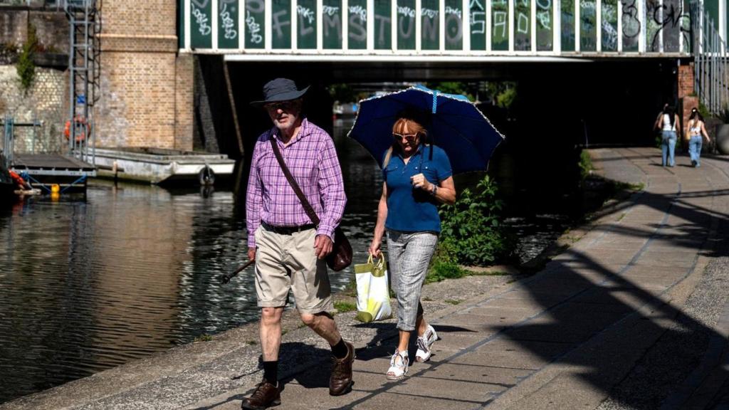 A man and woman walk beside Regent's Canal in London. The man wears a hat and the woman wears a hat shaped like an umbrella.
