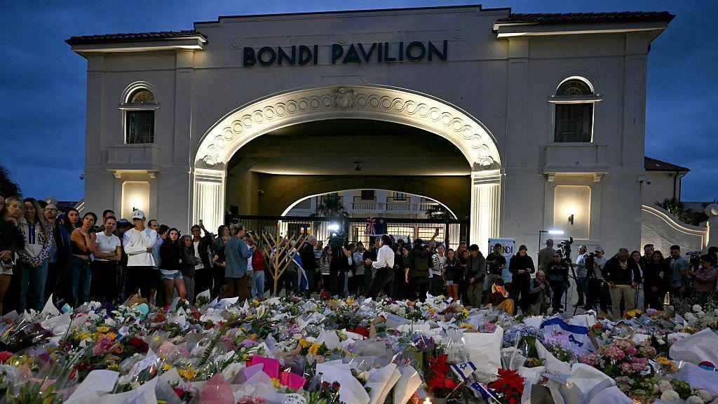 A large arched building in the background says "Bondi Pavilion". In the foreground, a large collection of tributes - made of of flowers, Israeli flags and other items - is laid on the floor in front of a crowd of people who have gathered. 