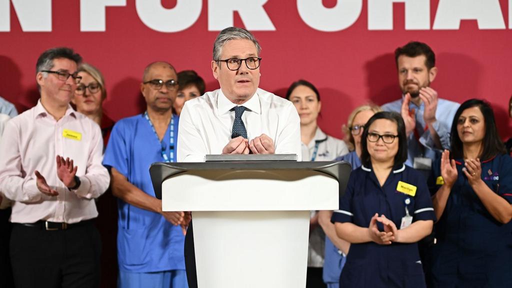 Starmer gestures while speaking with NHS staff in background