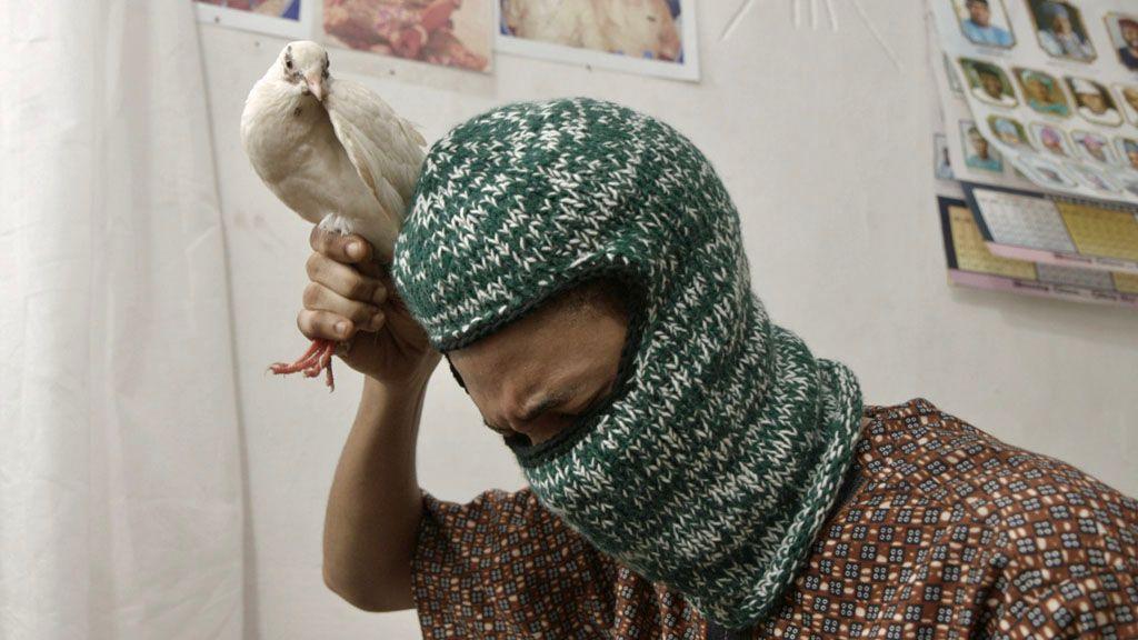Picture of a man leaning forward and looking down while holding a white dove by its legs, above his head.  The man is wearing a green woollen balaclava and a brown patterned short sleeve shirt.