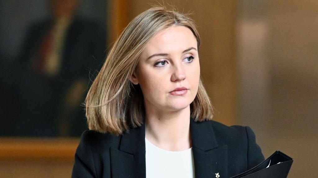 Candid image of Housing Secretary Màiri McAllan walking in the Scottish Parliament. She wears her hair in a stylish blonde bob and is looking off to the left as she carries her notes. She wears a black suit jacket and a white T-shirt.