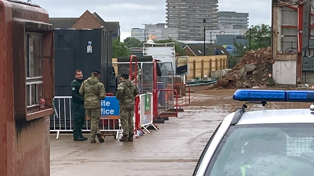 Army personnel speaking to a man next to a white barrier, with a construction site in the background