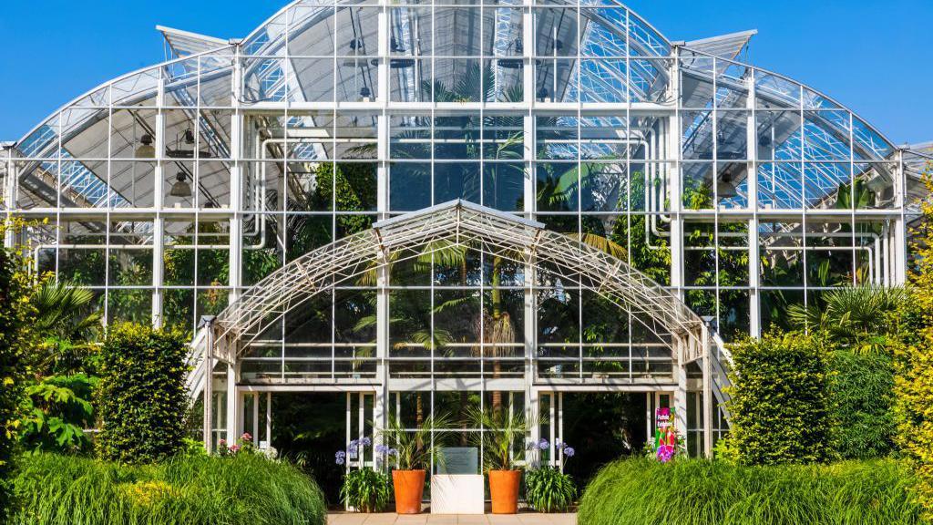 The glasshouse building at RHS Wisley. A front on view of the multi-story glass building. The sky is blue and there are lots of green bushes and plants around the building and inside. 