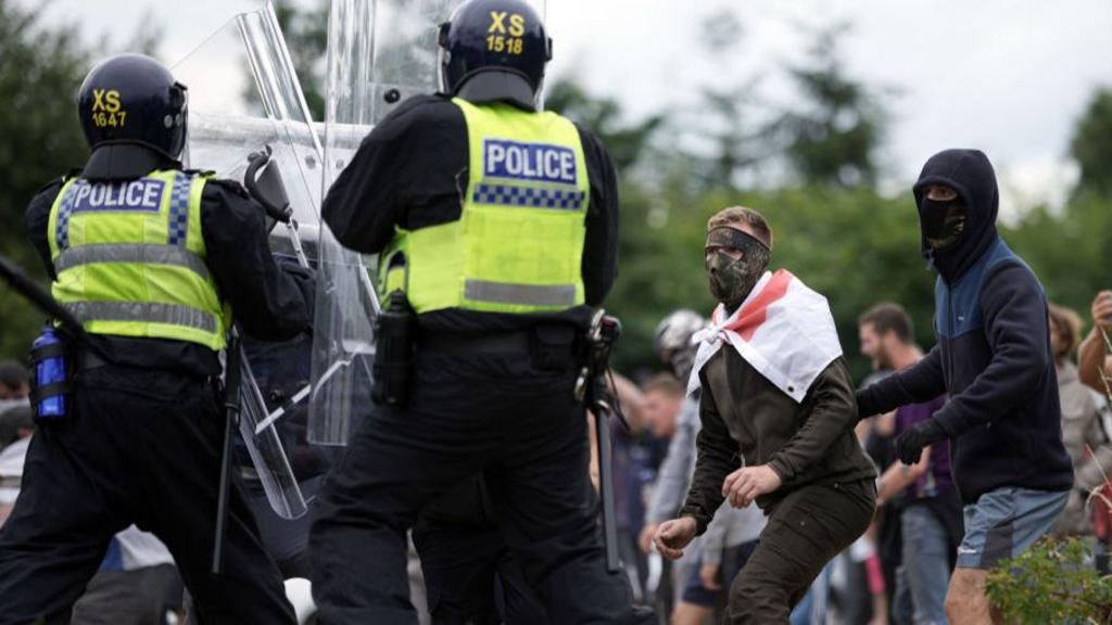 Two officers in riot gear with transparent rectangular shields face off against two masked men, one draped in a St George's Cross flag