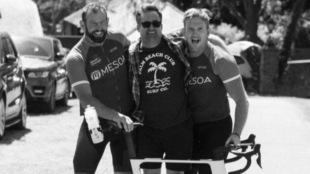 Tom Morris (middle), surrounded by two friends posing with a bike. All are smiling at the camera and the photo is in black and white, taken on a sunny day. 