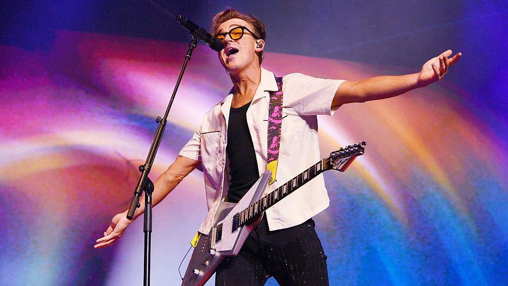 Tom Fletcher performing with an electric guitar and microphone on a stand. He is standing with his arms outstretched in front of a colourful background on a screen