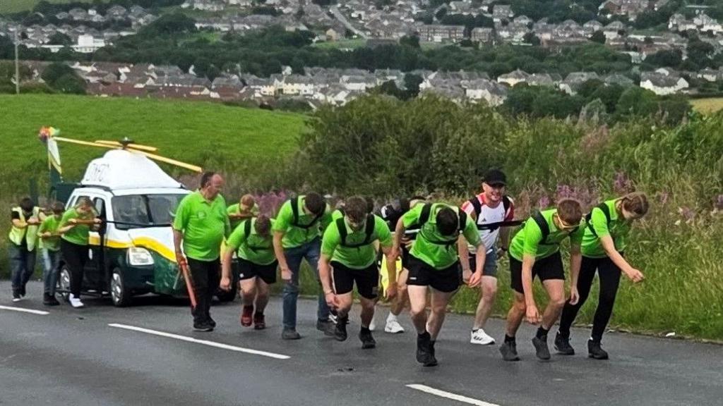 More than a dozen people are pulling a replica helicopter up a hill. A car has been painted green and white and fitted with a propeller to look like a Great North Air Ambulance helicopter. The group of young farmers are wearing bright green shirts and are attached to the replica via harnesses.