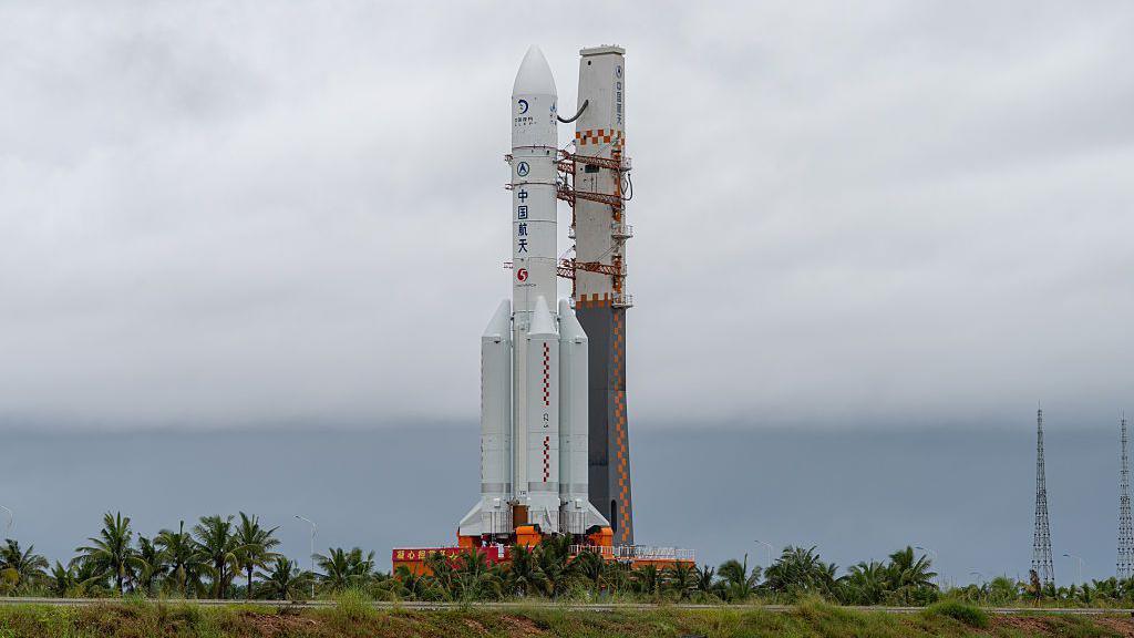 A white spacecraft standing. There are trees and grass around the bottom of it.