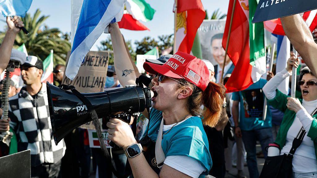 A woman in a Make America Great Again hat chants for regime change in Iran into a bullhorn during a protest in West LA in support of President Trump. A crowd behind her waves US, Israeli and Iranian flags.