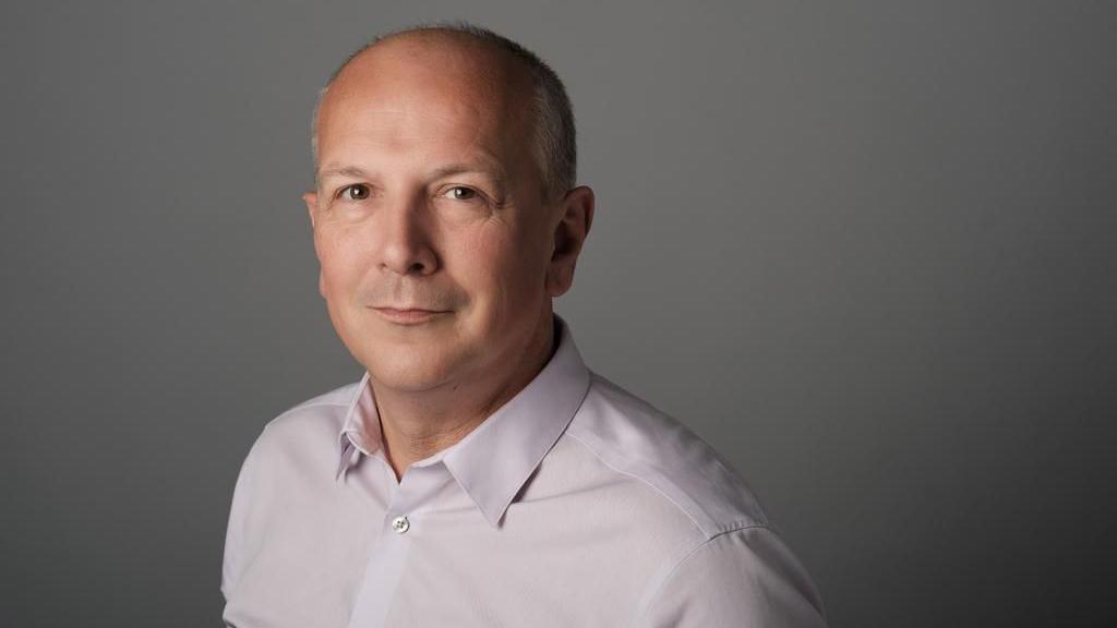 A professional-looking headshot of Simon Hewitt, a bald man wearing a light pink shirt. He's standing against a grey backdrop and looking straight at the camera.