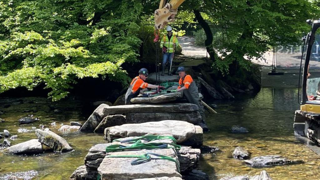 Workers in high-vis vests and hard hats use a digger to move huge slabs of stone crossing a shallow ford. Trees line the river bank.
