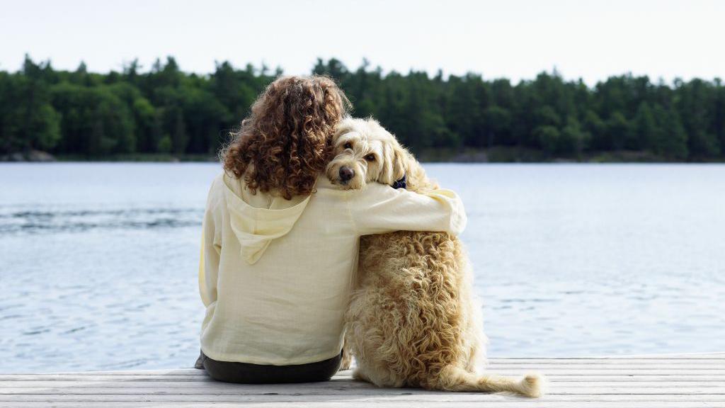 A woman sits on a jetty next to the water with her right arm around the neck of a sandy-coloured dog.  The dog has its head turned to the camera.