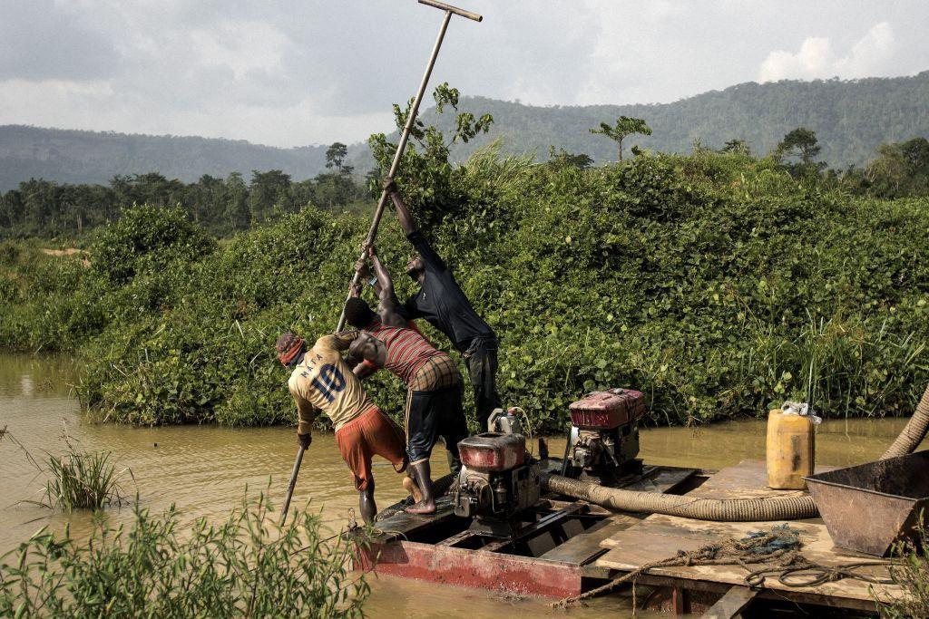 Three Illegal gold panners stand on a motorised pump to scrape the river bed as they look for specks of gold in the Kibi area in southern Ghana on 12 April 2017. The river is yellow-brown, with thick bushes on either side.