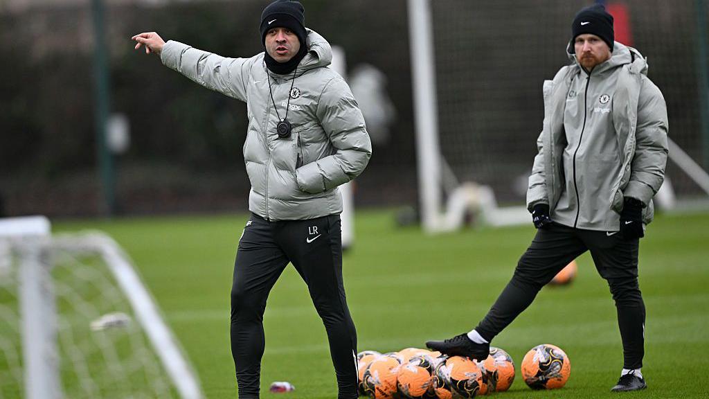 Head coach Liam Rosenior gesticulates to his players as he takes a Chelsea training session, with his arm outstretched. Fellow coach Calum McFarlane stands behind with a bunch of orange footballs, standing on one, both wearing Chelsea branded tracksuits of black beanie hats, great coats and black tracksuit trousers