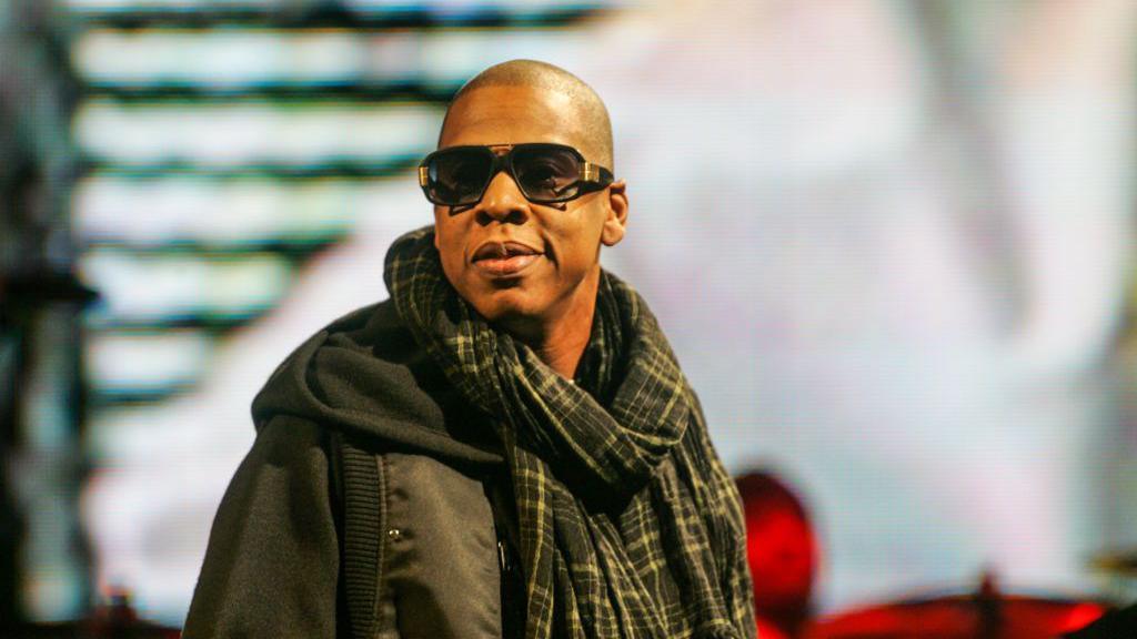 Jay Z on the Pyramid Stage. He is wearing dark sunglasses, a black and yellow checked scarf and a black hooded jacket.