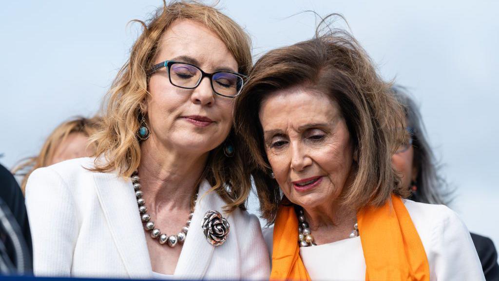 Gabrielle Giffords closes her eyes and rests her head against Nancy Pelosi's. Both are wearing white tops with silver jewellery, and Pelosi is wearing an orange scarf