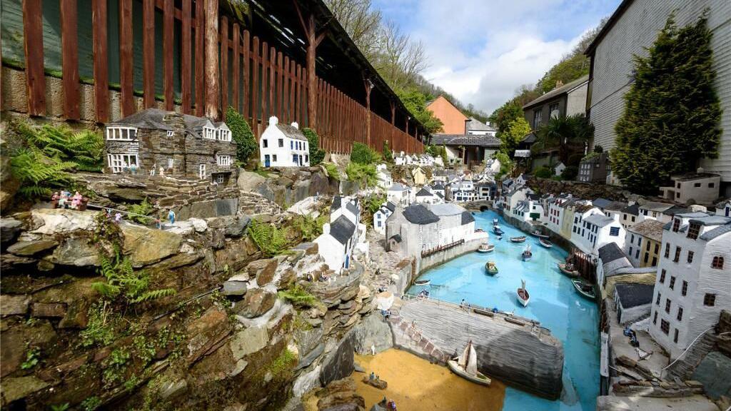 Some of Polperro Model Village, featuring various white-fronted model buildings along two banks of a narrow river, with the river having a harbour wall and a number of boats on it