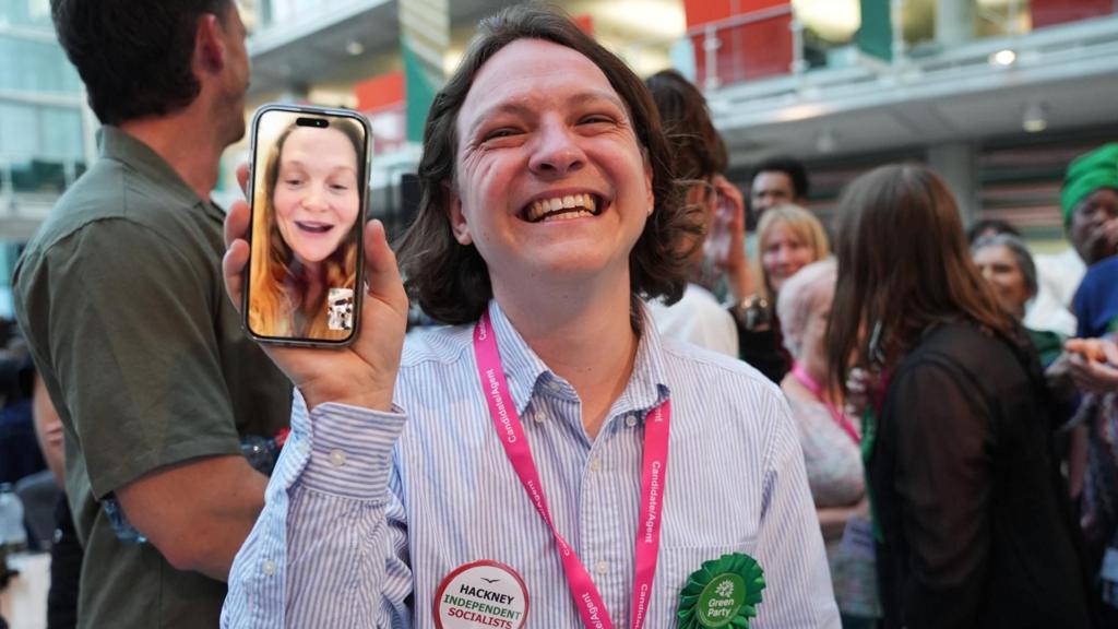 Green Party supporters congratulate candidate Florence Cullen-Davies - who is at home about to give birth and appears live via WhatsApp video on a mobile phone - after she is declared a winner in the 2026 London Borough of Hackne