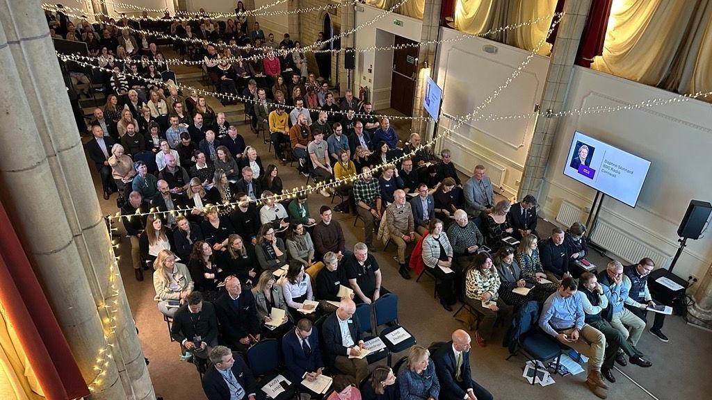 People sat on chairs in rows stretching across the room, which is light with columns and strings of lights above the people.