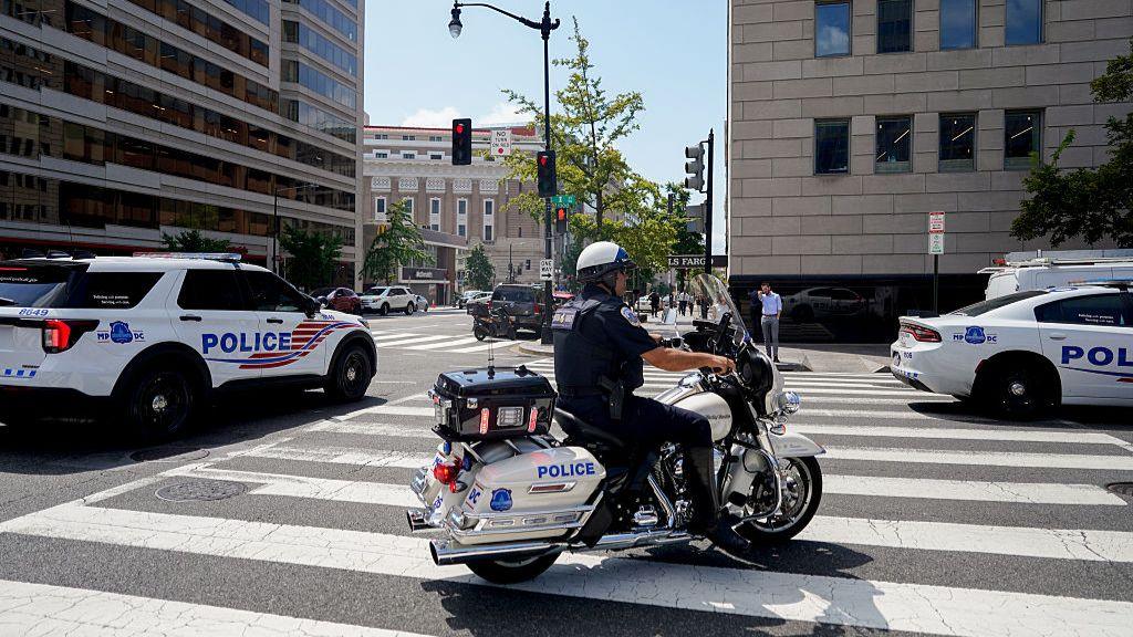 A Washington Police officer rides a police bike with two WPD police cars, one behind him and one parked ahead of him to his left, on a Washington street in the mid-morning sun