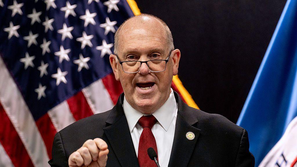 Tom Homan in dark suit with red tie in front of American flag, opens his mouth as he speaks and pinches his fingers 