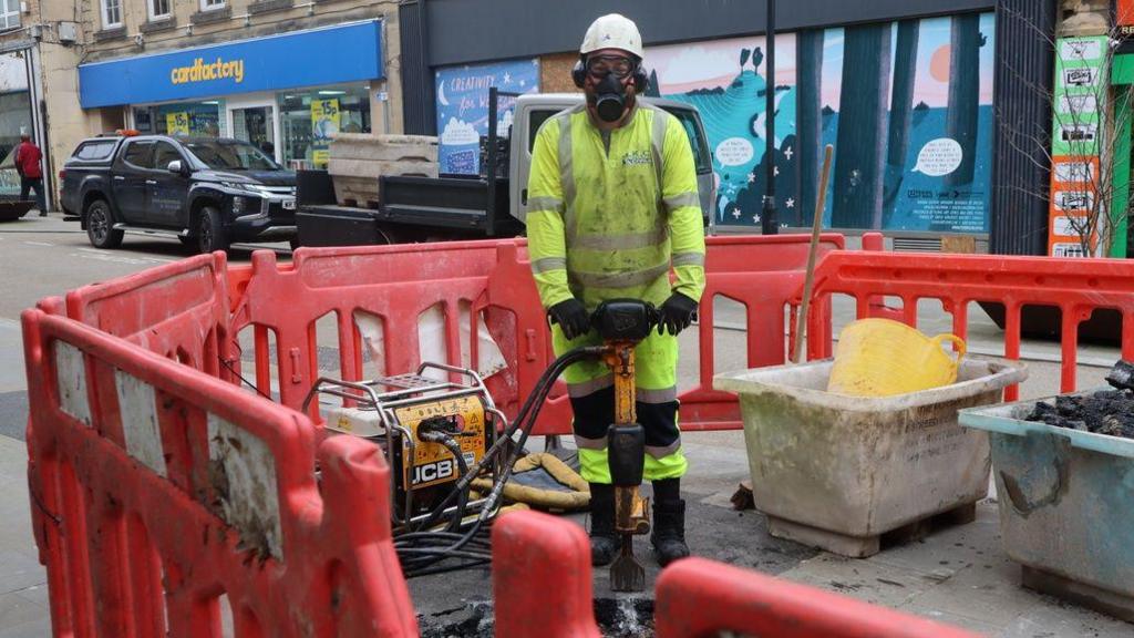 A workman operating a machine stands behind red fencing in the middle of the high street. He is wearing a bright yellow high-vis boiler suit, a face mask, goggles and a hard hat. 