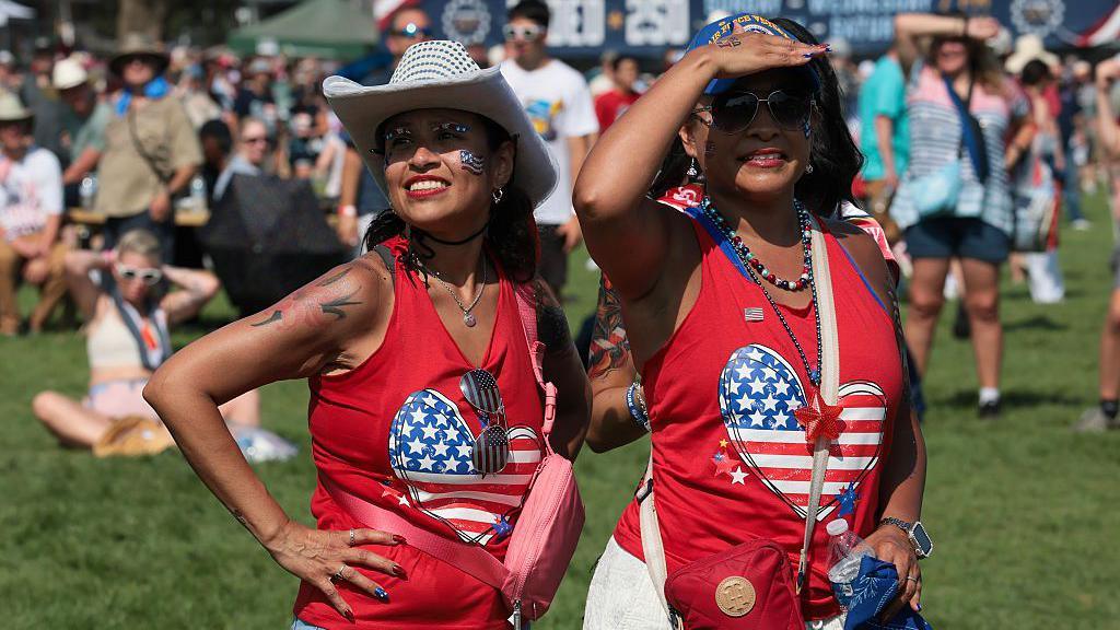 Two women in red tank tops with a heart-shaped US flag design wait on the lawn at the Great American State Fair in US. Both are wearing hats to shield them from the intense sunshine. Behind them, in the blurred background, are several people sitting on the grass