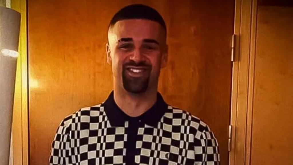 Tariq Evans is seen smiling at the camera wearing a black and white checked polo shirt. His black hair is short and he has a short beard and moustache.