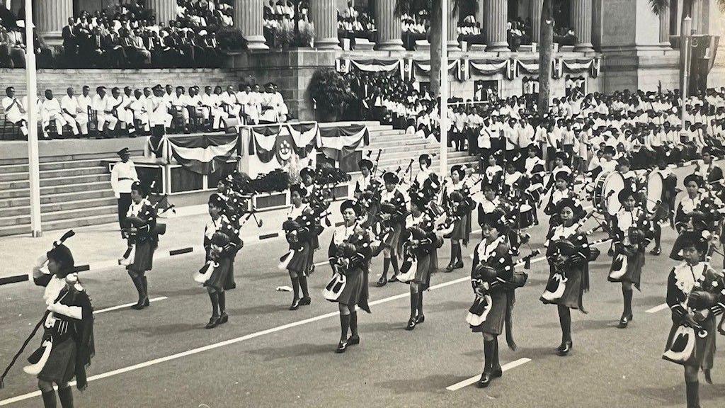 A black and white photo showing Singaporean girls in a marching band performing at an event. Officials, and the Singapore flag, can be seen in the background