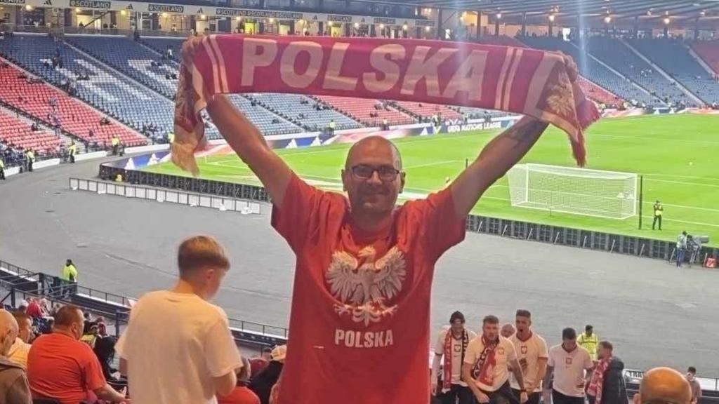 Jaroslaw Rossa, in a red Polska t-shirt, at a football match. He is holding a Polska scarf over his head.
