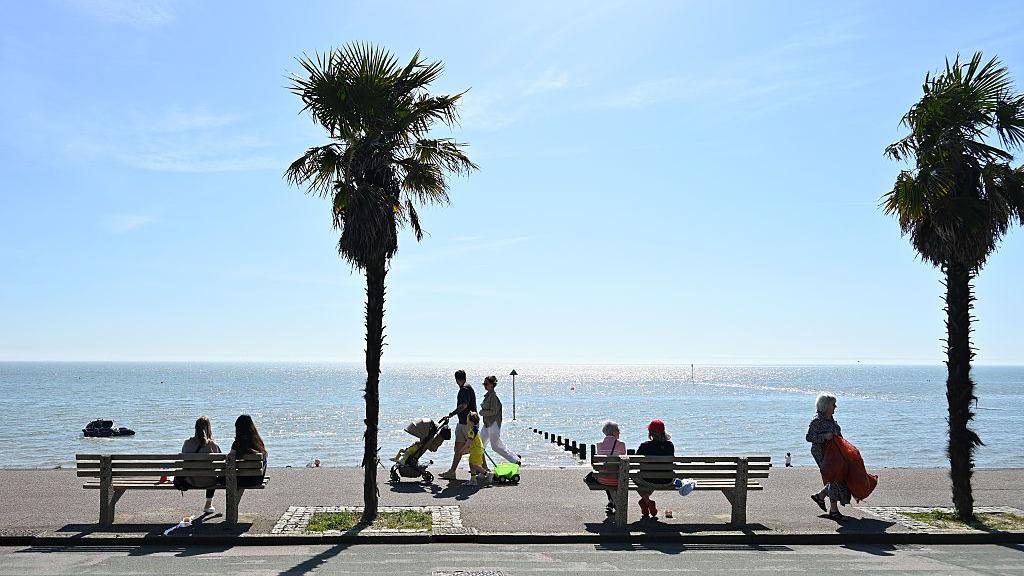 sunny scene of a coastal path where people are sitting on benches with other people walking along next to the glistening sea with blue skies overhead