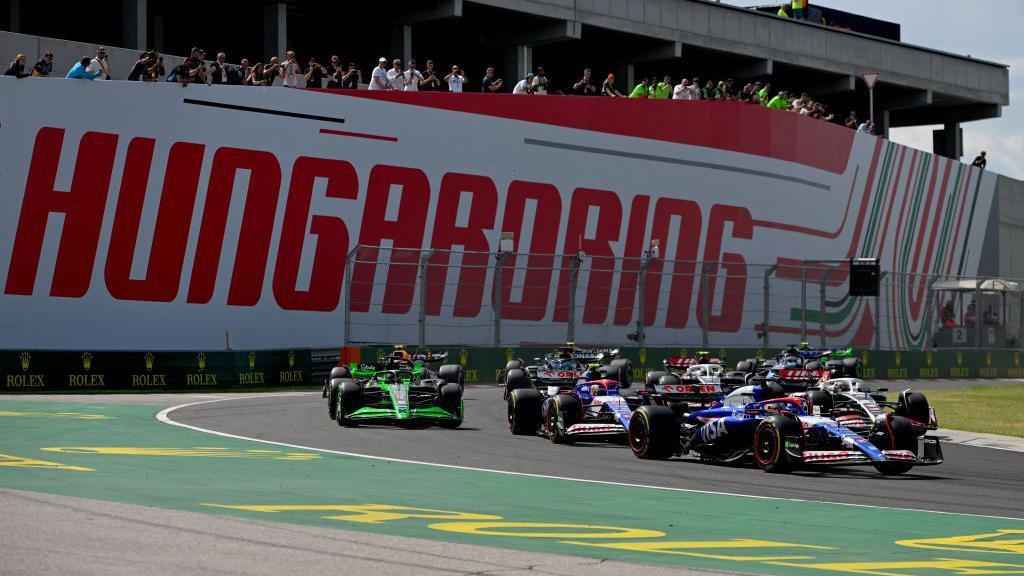 Formula One cars come through a corner during the 2024 Hungarian Grand Prix. There is a large wall in the background that says "HUNGARORING".