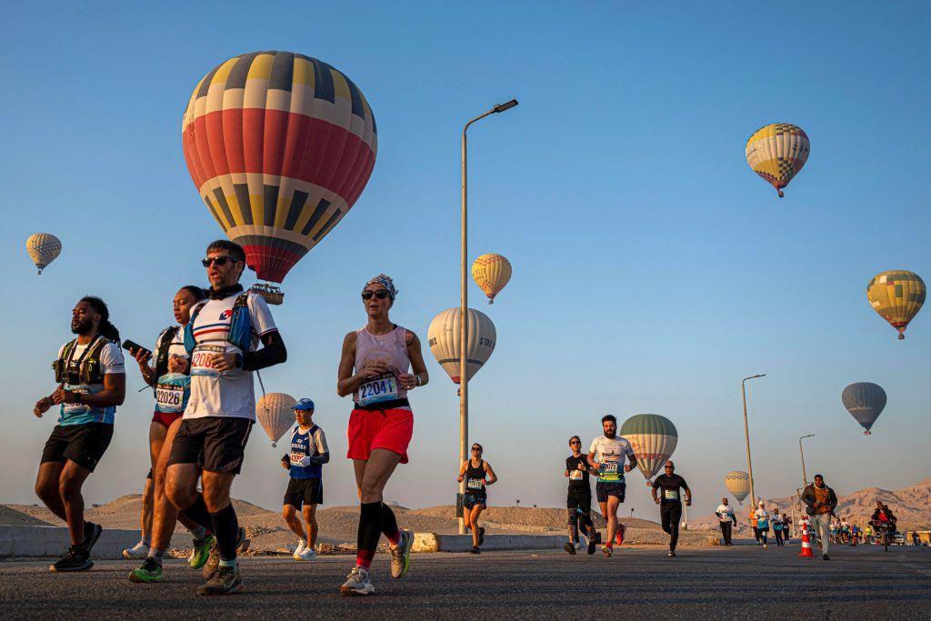 People in running gear and with numbers attached to their chests jog on a road. Hot-air balloons rise in the background.