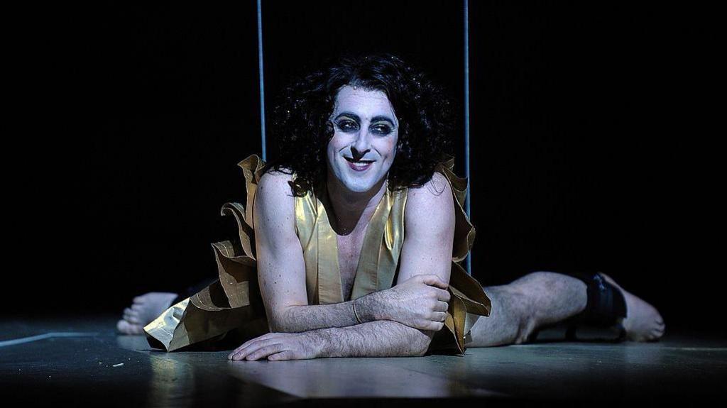 Alan Cumming on stage in white face make up with long black hair and a gold tunic. He is smiling and appears to he attached to wires like a puppet