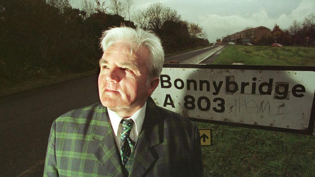 Billy Buchanan, a man with white hair, is wearing a green tartan suit and standing in front of a weathered road sign which reads Bonnybridge A803. Houses can be seen in the background. He is looking up into the air.