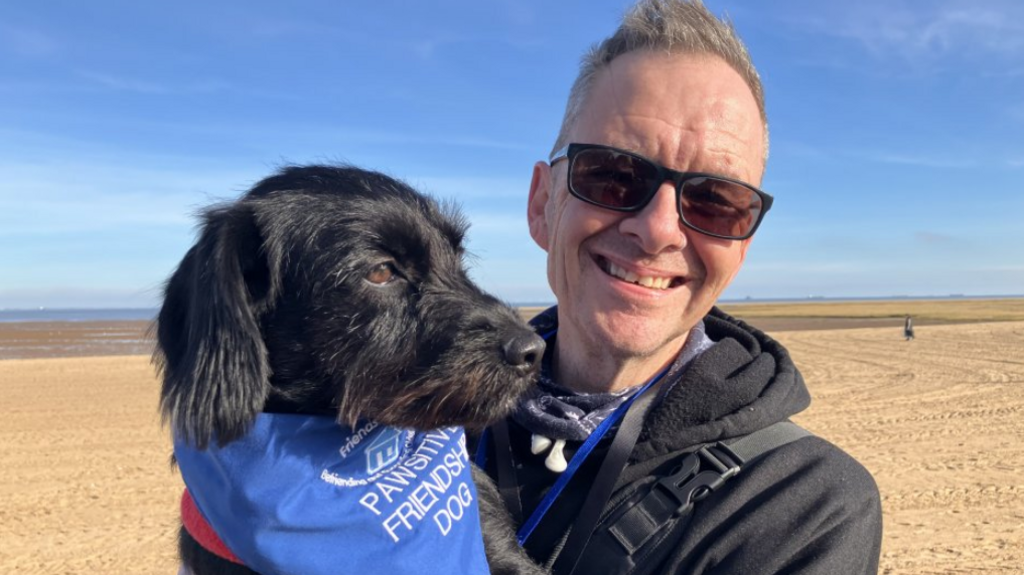 Smiling grey-haired man wearing sunglasses holding his pet dog, which has black fur and is wearing a blue bandana with the slogan "Pawsitive Friendship Dog". They are standing in front of a wide sandy beach and the sky is blue.
