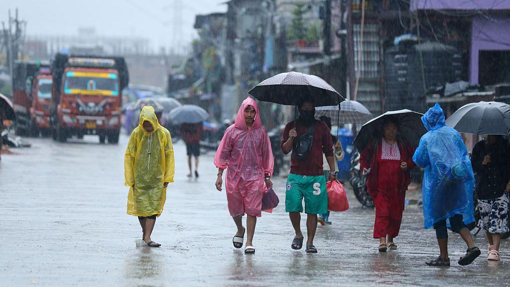 People in colourful raincoats, others with umbrellas, walking on a road in heavy rain