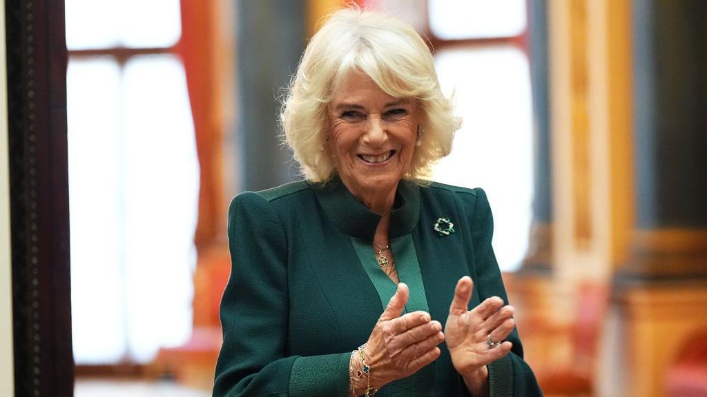 Queen Camilla during a reception for winners of the Queen's Commonwealth Essay Competition at Buckingham Palace. She is wearing green, with a green brooch on her left lapel. She appears to be mid-clap and is smiling