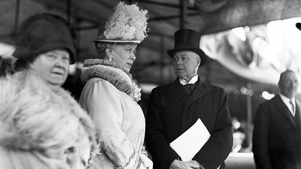 Black and white image of Queen Mary in an elaborate light coloured hat and coat standing beside a middle aged man in a black coat, white wing collar and black top hat.