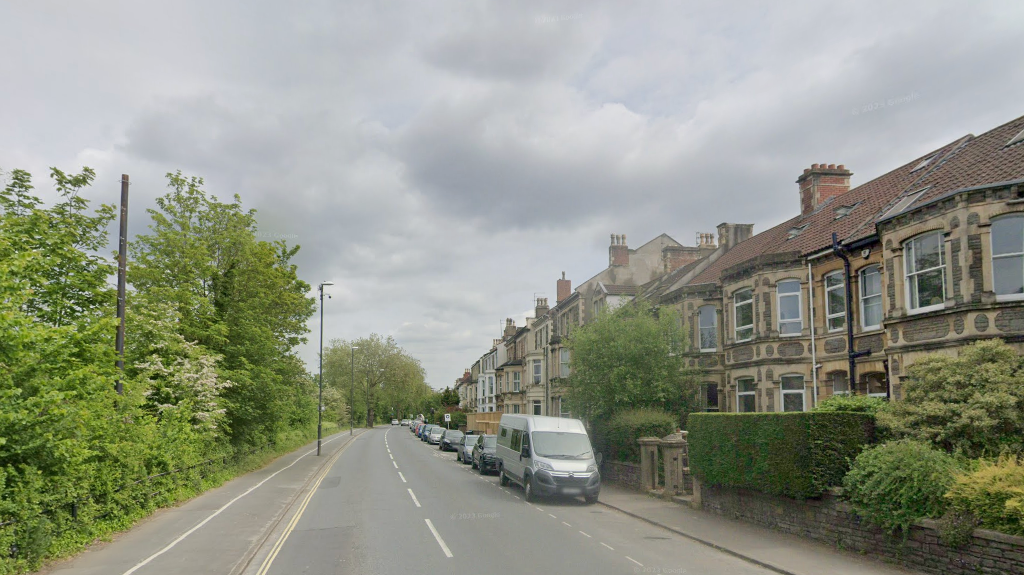 Coronation Road in Southville, Bristol. Houses are lining the street to the right, and trees and nature to the left. Cars and vans can be seen parked on the side of the road. 