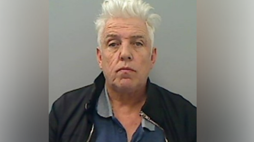 Peter Lamb with white hair that is sticking up looks straight at the camera. He is wearing a blue open t-shirt with a black zipped jacket. 