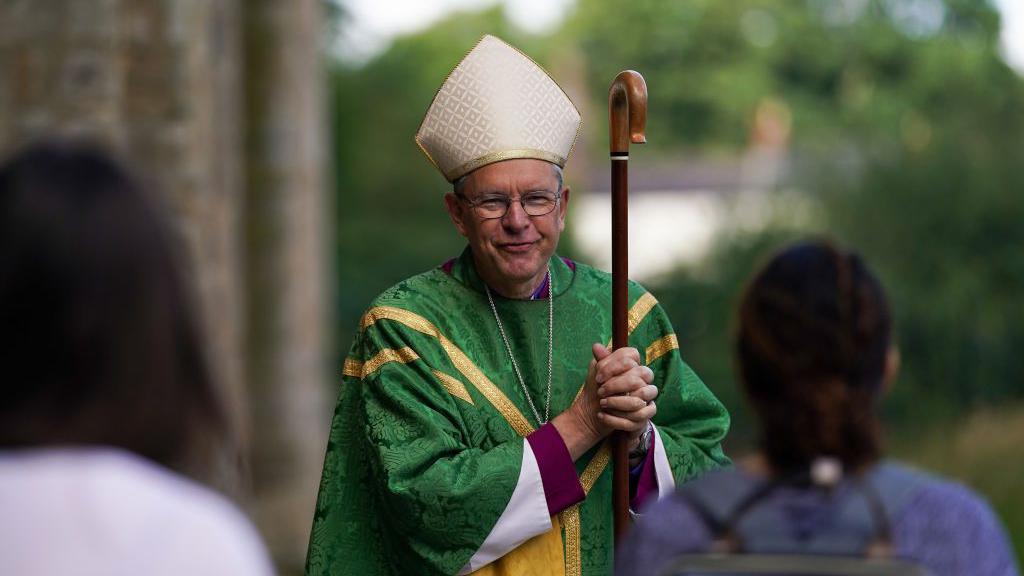 The Bishop of Durham, Paul Butler, speaking with members of the congregation after presiding over a Eucharist service in July 2020. He is wearing a green gown with gold detailing, and carrying a wooden stick. He has grey hair and glasses.