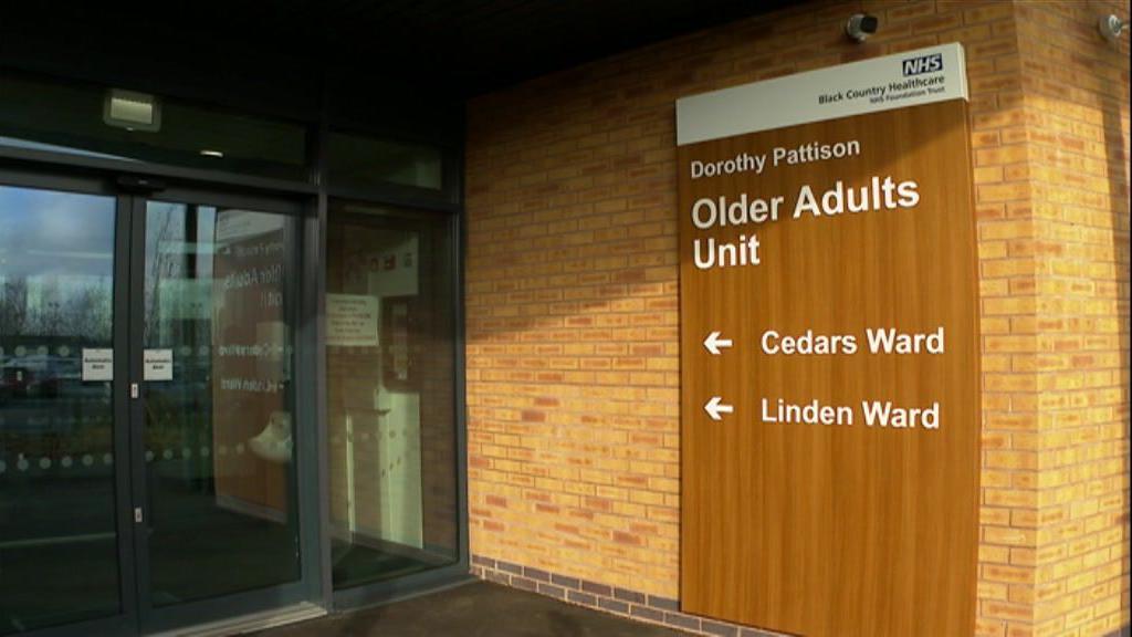 A wooden panel with the words Dorothy Pattinson Older Adults Unit, Cedars Ward and Linden Ward on a wall next to a glass sliding door.