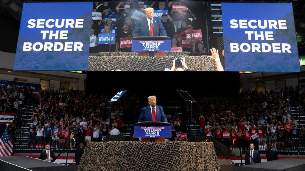 Donald Trump speaks at a rally in October in Arizona behind a podium. Two messages of "secure the border" are displayed flanking a big screen showing his speech. 