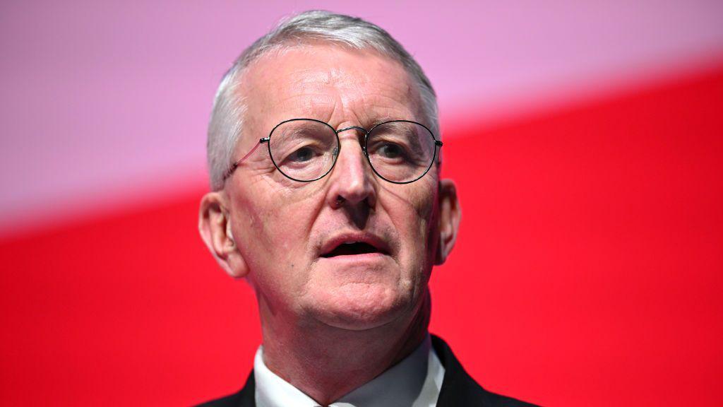 A close-up shot of Hilary Benn in front of a red background. He has short grey hair and round-rimmed glasses.