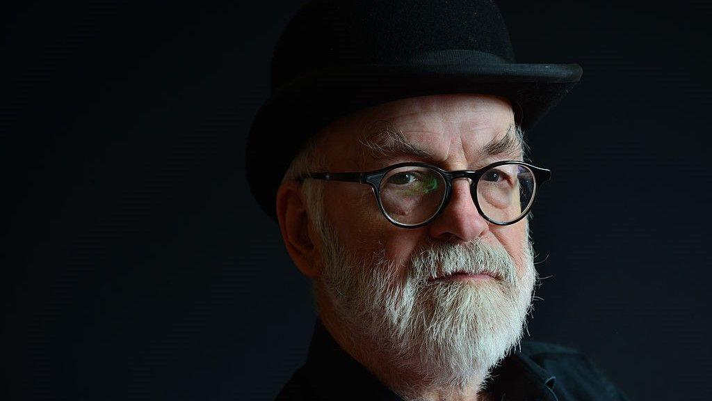 Portrait of Terry Pratchett, who is wearing a black shirt and black brimmed hat, in front of a plain black background. He has a white/grey beard and is wearing black glasses.