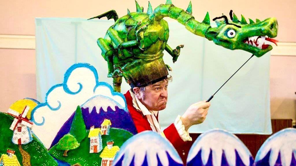 Paul with a green dragon puppet that rests on his head like a hat, using a stick to manoeuvre the rest of its body in a home made set.