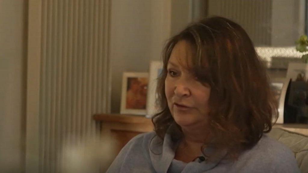 Joanne Gardiner is sitting on a sofa in her home. There are family photos in the background. She has shoulder-length curly brown hair.