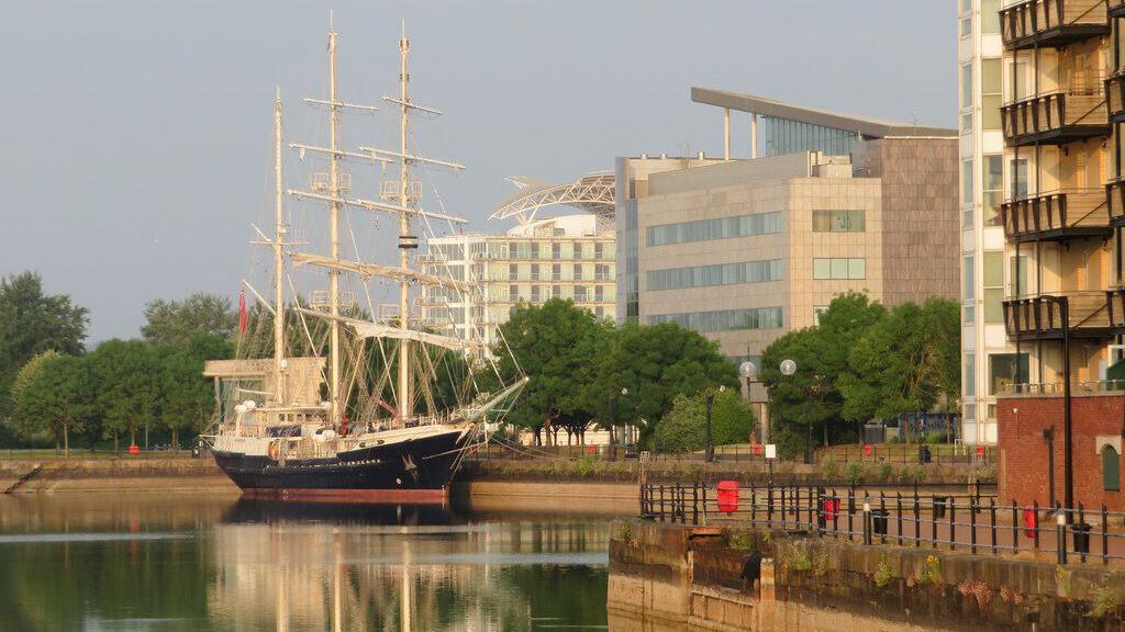 A schooner with a blue and red hull and white decking and masts is moored alongside Britannia Quay in Cardiff Bay.