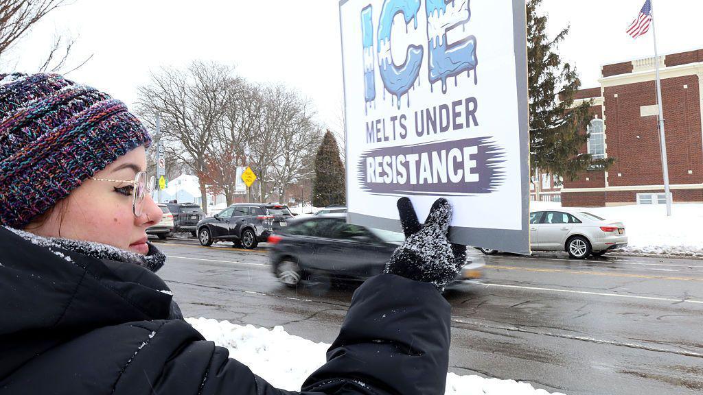 Protester, in profile, stands on a snowy roadside, wearing a tuque, glasses, and black winter coat and gloves, holds a sign that reads: "ICE melts under resistance."